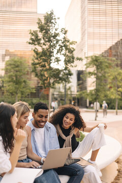 Nice young interracial students use laptop after class sitting outdoors. Guy with girls wear casual clothes in spring. Concept of modern education