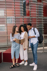 Full length of three young diverse classmates checking their homework before lessons outside. Brunette guy and girls wear casual clothes. Knowledge concept
