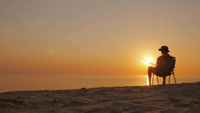 Silhouette of a lonely man sitting on a chair by the lake and catching fish at sunset