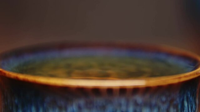SIngle big drop falling into a beautiful blue and orange ceramic teacup filled to the brim with green tea, ripples forming, steam rises. Extreme macro closeup of hot beverage