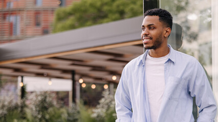 Fototapeta premium Handsome young african guy is looking towards city relaxing outdoors. Brunette man with stubble wears t-shirt and shirt. Lifestyle concept