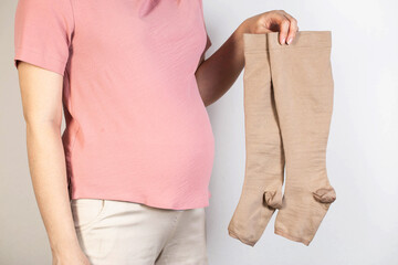 A pregnant girl in a pink tank top holds medical compression stockings for varicose veins in her...