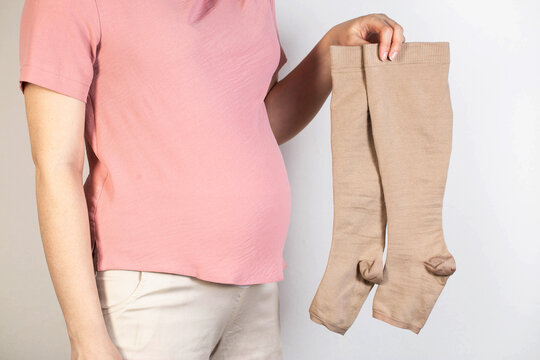 A pregnant girl in a pink tank top holds medical compression stockings for varicose veins in her legs. White background. Pregnancy and varicose veins, phlebology