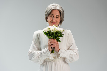 joyful middle aged bride in white wedding dress holding bouquet and smelling flowers isolated on grey