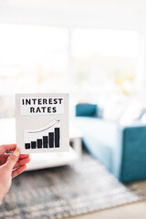 property affordabilikty and home loan payment increases, hand holding interest rates sign with graph showing stats going up in front of living room