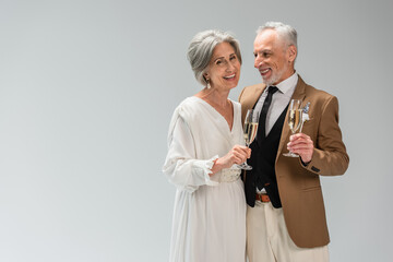 middle aged groom and happy bride in wedding dress holding glasses of champagne isolated on grey