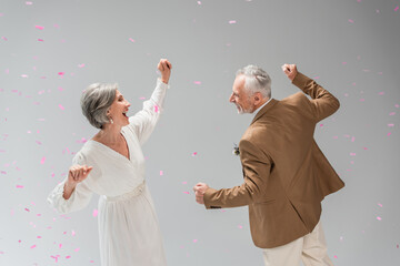 cheerful middle aged groom and happy bride in white dress dancing under falling confetti on grey