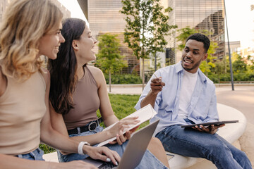 Diverse young classmates having fun chatting and using modern technology on street. Girls and guy spending their leisure time studying. Student concept