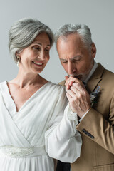 middle aged man in suit kissing hand of happy bride in white wedding dress isolated on grey