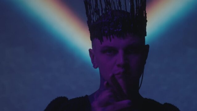 Tilt down shot of gothic man with gothic crown and black painted nails dancing in dark studio with rainbow light projection