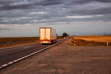 A truck with a trailer transports cargo in the summer against the backdrop of sunset and beautiful...