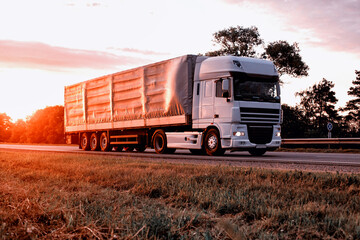 A truck with a tilt semi-trailer transports cargo against the backdrop of a sunset in the summer. Sharing economy concept in freight logistics. Modernization of logistics companies