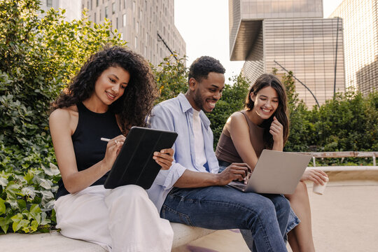 Smiling young interracial students spend time together working on laptop and tablet outdoors. Brunette guy and girls wear casual clothes. Concept of learning