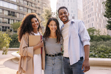 Three positive young interracial students look at camera standing outdoors. Brunettes guy and girls wear casual clothes for walk. Real emotions concept