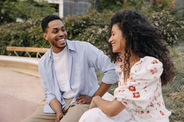 Cheerful african young couple are relaxing outdoors enjoying each other. Brunette girl and guy are wearing shirt with blouse. Happy day concept