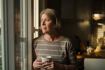 mature disturbed woman looking through the window while standing in her kitchen