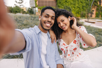 Beautiful young african couple take photo of themselves using headphones sitting in nature. Brunette guy wears shirt, girl wears blouse. Relaxed lifestyle, concept