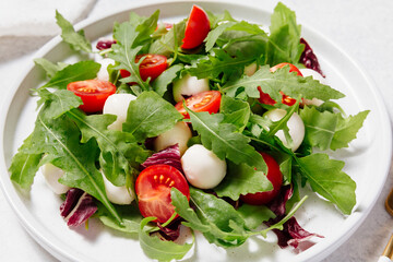 Fresh green salad made with mozzarella cheese, cherry tomatoes, arugula and olive oil on white stone table background. Healthy food and diet concept. Flat lay, top view, copy space.
