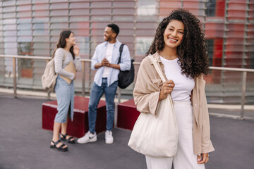 Pretty young dark-skinned brunette girl poses for camera standing near university during day. In background, interracial students are chatting with each other. Lifestyle concept