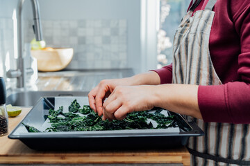 Close up woman laying out teared curly green kale leaves on a baking sheet on the kitchen. Cooking kale chips. Healthy eating, dieting. Step by step cooking. Selective focus, copy space.