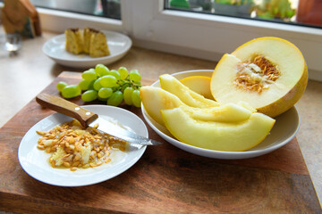 sliced melon with seeds on a white plate, a kitchen knife and green grapes on a wooden board, windowsill, the concept of fresh fruit and healthy eating