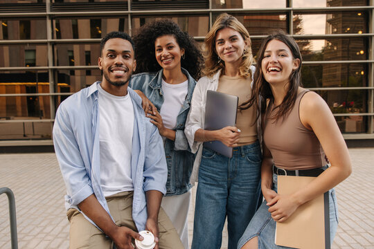 Happy young interracial students guy and girl looking at camera standing outdoors. People wear casual clothes and hold study items. Lifestyle concept