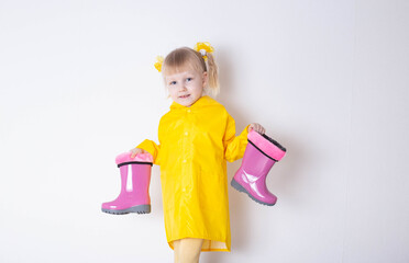 A little caucasian girl aged 3 years in a yellow raincoat holds pink rubber boots in her hands on a white background, isolate. The concept of bad weather and autumn.