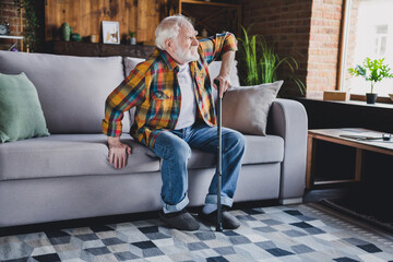 Photo of stressed depressed retired man dressed plaid shirt holding nursery cane trying getting up couch indoors apartment