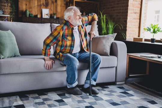 Photo of stressed depressed retired man dressed plaid shirt holding nursery cane trying getting up couch indoors apartment