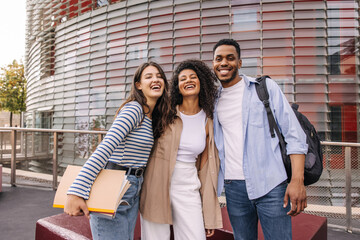 Joyful young interracial guy and girls posing looking at camera standing outdoors. Brunette students wear casual clothes. Good mood concept