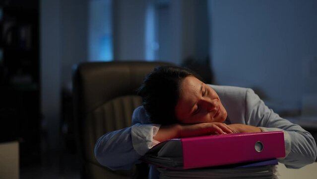 Exhausted woman taking a nap leaning head on document folders in office. Portrait of tired Caucasian female employee falling asleep closing eyes sitting indoors in the evening. Tiredness and business