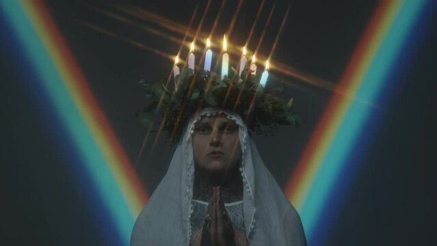 Zoom in shot of man with candle lit wreath on his head posing for camera with hands folded in dark studio with rainbow light projection in background