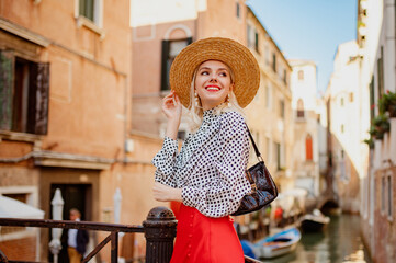 Happy smiling fashionable blonde woman wearing summer straw hat, polka dot blouse, red skirt, posing on the bridge in Venice. Fashion, travel, lifestyle conception. Copy, empty space for text