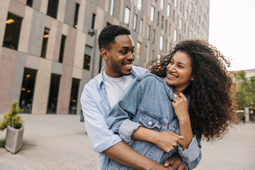 Beautiful young african couple looking at each other hugging standing outdoors. Brunette guy and girl wear casual clothes. Concept of enjoyment