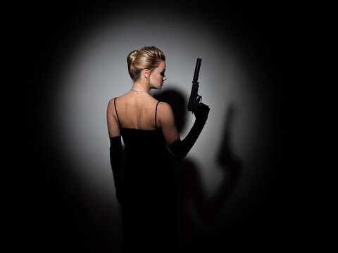 A femme fatale is aiming a silenced pistol, a spy or an undercover agent, a portrait in a spot of light