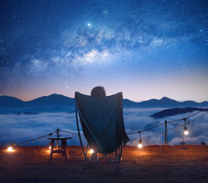 woman in camping chair looks at starry sky and sunrise in mountains.
