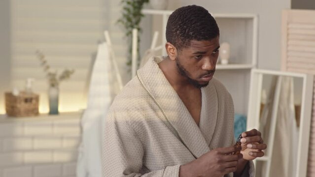Medium slowmo of handsome young Black man in bathrobe applying and rubbing nourishing serum on his face after morning shower, doing skincare procedures in front of mirror in bathroom
