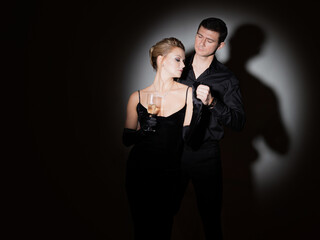 An attractive couple in black. Stylish and elegant young man and woman in the spotlight. Beautiful blonde in a long dress with a glass in her hands