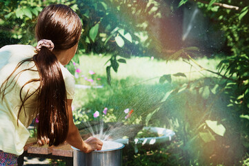 Brunette girl with long hair plays with a drinking fountain in the park in summer. Rainbow in...