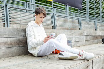 Teenage boy uses smartphone and wireless headphones during break on basketball court.