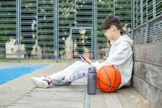 Teenage boy uses smartphone and wireless headphones during break on basketball court.