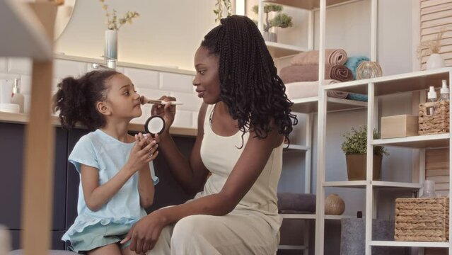 Medium slowmo of young beautiful African American woman and her pretty 6 year old daughter doing makeup to each other in bathroom