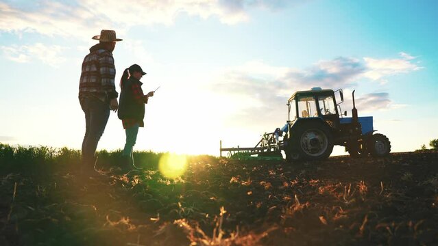 Silhouette farmer woman and man engineer with digital tablet walking in field in rays of sunset on field. Teamwork in agribusiness. Farmers inspect their rural surroundings. Summer walk in nature.