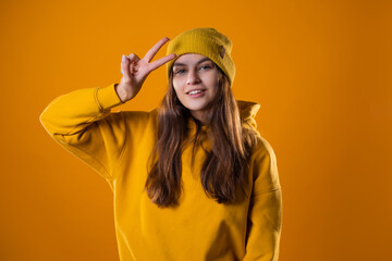 Stylish cheerful young brunette in a yellow hoodie and hats shows a victory gesture with two fingers up, on a yellow background
