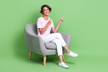 Full length photo of pretty young girl sit chair fingers point empty space wear stylish striped...