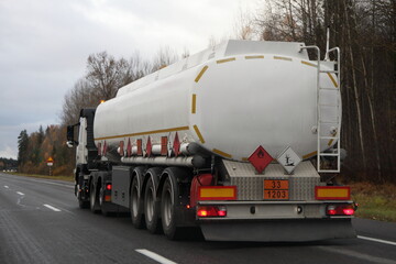 Petrol transportation, white fuel tanker truck with ADR sign 33 1203 on suburban empty asphalted road at autumn evening on forest background, flammable liquid goods transportation