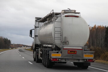 Big semi truck with barrel for liquid food goods drive on suburban highway road at autumn evening in perspective, back side view