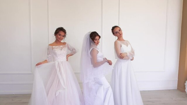 Three beautiful girls in wedding dresses pose for the photographer. Charming young brunette bride on a light isolated background in a white dress. Space for copy. UHD 4K.