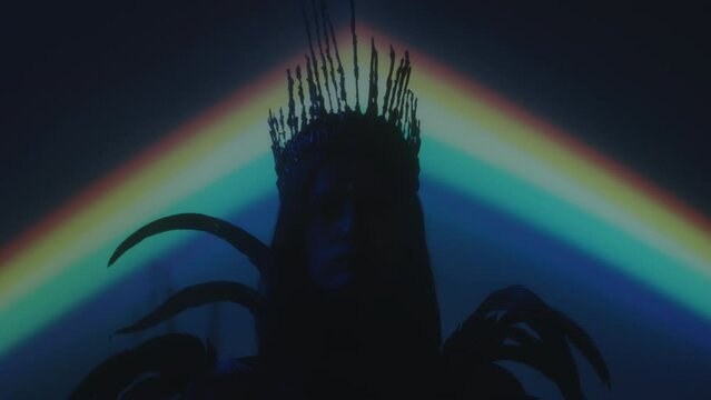 Zoom out portrait shot of woman wearing gothic crown and feather dress posing for camera in dark studio with blue neon light and rainbow projection