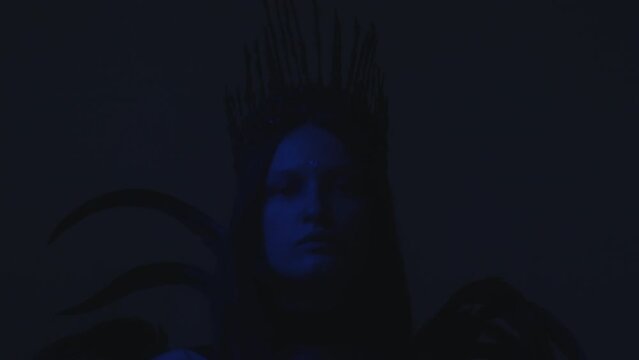 Zoom out shot of woman with shining bindi wearing gothic crown and feather dress posing for camera in dark studio with neon light and rainbow projection